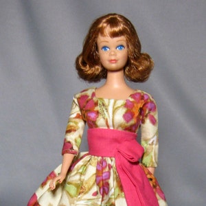 May include: A vintage doll wearing a floral print dress with a pink sash. The doll has red hair and blue eyes.