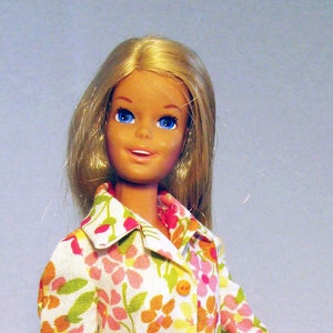 May include: A blonde doll wearing a yellow skirt with pink buttons and a floral print blouse. The doll is wearing yellow shoes and is standing on a clear plastic stand.