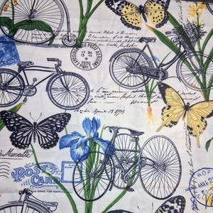 May include: White fabric with a repeating pattern of bicycles, butterflies, postage stamps, and handwritten script. The bicycles are black and white, and the butterflies are yellow and black. Blue iris flowers and green foliage are also present.