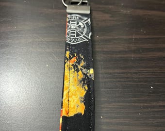 Könnte beinhalten: Ein Schlüsselanhänger aus Stoff mit schwarzem Hintergrund und einem aufgedruckten Feuer- und Flammenmuster. Der obere Teil zeigt ein weißes Malteserkreuz-Emblem mit dem Text FIRE RESCUE. Ein silberner Metallverschluss und ein Schlüsselring sind befestigt.