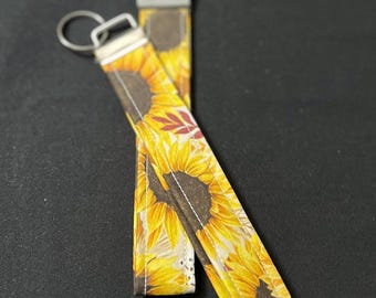 Könnte beinhalten: Zwei Schlüsselanhänger mit Sonnenblumenmuster und silbernen Schlüsselringen. Die Schlüsselanhänger zeigen ein leuchtend gelbes Sonnenblumendesign mit braunen Zentren und grünen Blättern. Der Hintergrund ist einfarbig schwarz.