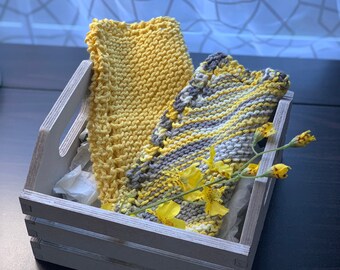 Könnte beinhalten: Eine graue Holzkiste enthält zwei gestrickte Waschlappen. Einer ist einfarbig gelb, der andere eine Mischung aus Gelb, Grau und Weiß. Gelbe Blumen sind obenauf gelegt. Die Kiste steht auf einer dunklen Oberfläche.