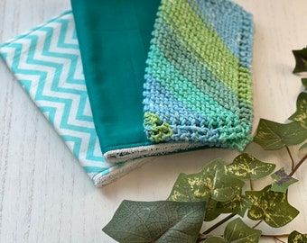 Könnte beinhalten: Drei handgemachte Waschlappen. Einer ist türkis, einer ist weiß mit einem türkisfarbenen Zickzackmuster, und der dritte ist ein gestrickter Waschlappen mit blauen und grünen Streifen. Die Waschlappen liegen auf einer weißen Holzoberfläche mit grünem Efeu.
