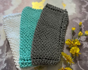 Könnte beinhalten: Drei gestrickte Waschlappen in Weiß, Türkis und Grau sind aufeinander gestapelt. Die Waschlappen bestehen aus einem strukturierten Garn und haben eine rechteckige Form. Ein Zweig gelber Blumen mit braunen Flecken befindet sich im Hintergrund.