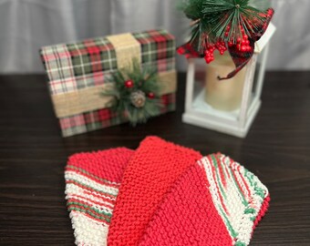 Könnte beinhalten: Drei gestrickte Waschlappen in Rot, Weiß und Grün. Zwei sind einfarbig rot, und einer hat rot, weiß und grüne Streifen. Eine karierte Geschenkbox mit einem Tannenzapfen und eine weiße Laterne mit einer Kerze und Grünzeug befinden sich im Hintergrund.