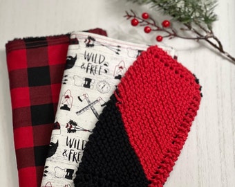 Könnte beinhalten: Ein Stapel aus drei Stoffartikeln. Der erste hat ein rot-schwarzes Büffelkaro-Muster. Der zweite ist weiß mit einem "WILD & FREE"-Druck mit Äxten und anderen Grafiken. Der dritte ist ein rot-schwarzer Strickartikel.