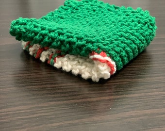 Könnte beinhalten: Ein gefalteter, gestrickter Waschlappen mit einer grünen oberen Schicht und einer weißen unteren Schicht. Die Kanten des Waschlappens sind rot eingefasst. Der Waschlappen besteht aus einem dicken, strukturierten Garn.