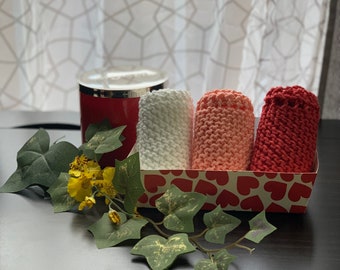 Könnte beinhalten: Ein dekoratives Geschenkset mit drei gerollten, gestrickten Waschlappen in Weiß, Pfirsich und Rot, präsentiert in einer herzförmigen Box. Eine rote Kerze und künstliches Grünzeug ergänzen das Arrangement.