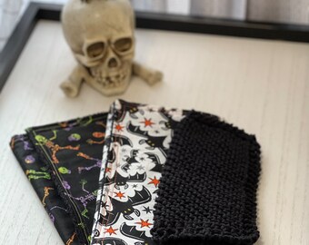 Könnte beinhalten: Halloween-Dekorationsartikel. Enthält eine schwarze Krähenfigur auf einem Totenkopf und gefaltete Stoffservietten mit schwarz-weißem Muster mit Halloween-Motiven wie Skeletten, Fledermäusen und Sternen. Ein schwarzer Strickartikel ist ebenfalls vorhanden.