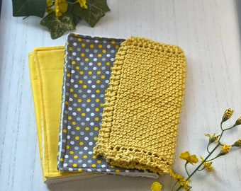 Könnte beinhalten: Ein Stapel handgefertigter Waschlappen in Gelb und Grau. Die Waschlappen weisen verschiedene Muster auf, darunter einfarbiges Gelb, Grau mit weißen und gelben Punkten und ein gestrickter gelber Waschlappen. Gelbe Blumen und grüne Blätter sind im Hintergrund angeordnet.
