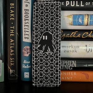 Pode incluir: Um marcador de livros de tecido preto e branco com um padrão repetido de pequenas figuras de fantasmas e um design de fantasma maior. O marcador de livros é colocado em frente a uma pilha de livros.
