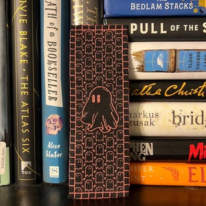 May include: A black bookmark with a pink ghost design. The bookmark has a repeating pattern of small ghosts and a larger ghost in the center. The edges are outlined in pink. The bookmark is standing in front of a stack of books.
