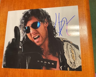 May include: A glossy photograph featuring a person with long dark hair wearing dark sunglasses and a black leather jacket, pointing upward with a gloved hand. A large metallic championship belt is visible on the shoulder, and a blue ink signature is present.