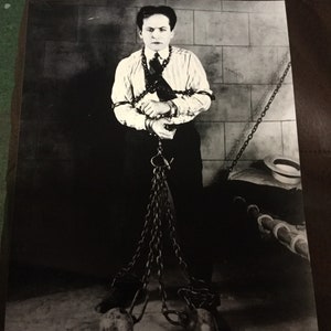 Può includere: Una fotografia in bianco e nero di un uomo con una camicia bianca e pantaloni scuri, con le mani legate da catene e attaccate a pesanti sfere di metallo. L'uomo è in piedi di fronte a un muro di mattoni.