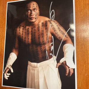May include: A glossy photograph featuring a muscular individual with extensive black ink calligraphy tattoos covering their face, chest, and arms. The individual wears white wrist wraps and white trousers, with a white handwritten signature overlay.