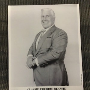 May include: Black and white photo of a man in a suit. He is standing with his arms crossed and looking at the camera. The photo is captioned "CLASSSIE FREDDIE BLASSIE" and "1987 Titan Sports, Inc. For Newspaper Use Only."