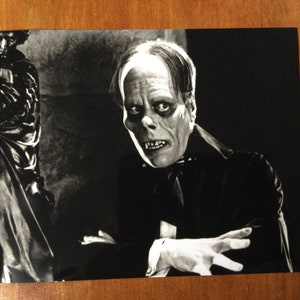 May include: Black and white photograph of a man with a mask covering his face. The man is wearing a dark suit and has a serious expression.