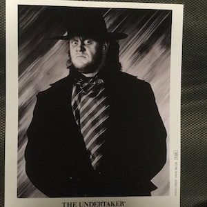 Puede incluir: Foto en blanco y negro de The Undertaker, un luchador profesional, con un sombrero negro y un traje con una corbata a rayas. La foto es de 1990 e incluye el texto "The Undertaker" y "World Wrestling Federation".