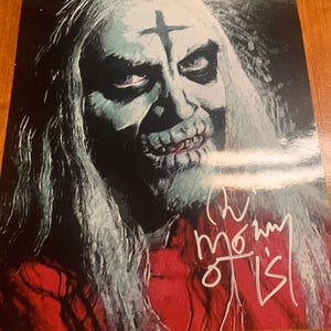 Peut inclure: Une impression rectangulaire représentant le portrait d'une silhouette pâle aux longs cheveux avec une croix sombre sur le front, vêtue d'un vêtement rouge. L'image comporte une signature manuscrite blanche en bas à droite, lisible Rob Zombie.