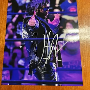 May include: A signed photograph of a wrestler in a wrestling ring. The wrestler is wearing a black leather jacket, black pants, and sunglasses. The wrestler is pointing with both hands. The background shows a crowd of people.