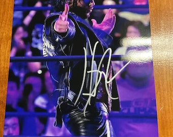 May include: A signed photograph of a wrestler in a wrestling ring. The wrestler is wearing a black leather jacket, black pants, and sunglasses. The wrestler is pointing with both hands. The background shows a crowd of people.