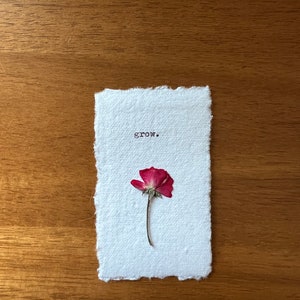 Puede incluir: Una tarjeta de papel blanco hecha a mano con la palabra "grow." impresa en negro. Una rosa roja seca está fijada debajo del texto. La tarjeta tiene un borde irregular y está sobre un fondo de madera.