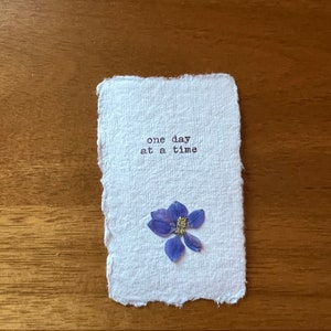 Puede incluir: Una tarjeta de papel blanco hecha a mano con la frase "one day at a time" impresa en negro. Una pequeña flor azul seca está adherida a la tarjeta. La tarjeta descansa sobre una superficie de madera.