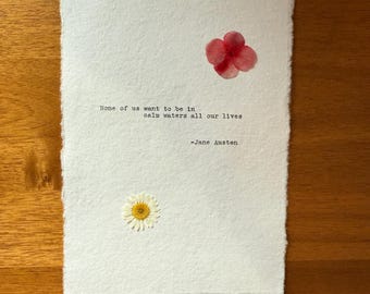 Könnte beinhalten: Ein weißes Papier mit einem Zitat von Jane Austen, verziert mit einer roten Blume und einem Gänseblümchen. Das Zitat lautet: „Keiner von uns will in ruhigen Gewässern sein, unser ganzes Leben lang.“ Das Papier hat einen strukturierten, gerissenen Rand, vor einem Holzgrund.