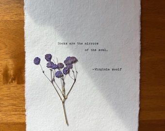 Könnte beinhalten: Texturiertes weißes Papier mit dem Zitat "Bücher sind die Spiegel der Seele." und dem Namen des Autors, Virginia Woolf, in Schwarz gedruckt. Ein Zweig getrockneter lila Blumen ist auf dem Papier befestigt.