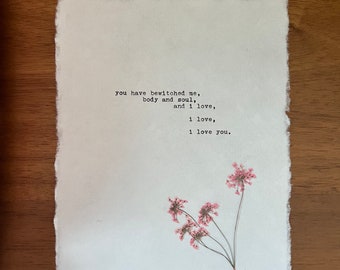 May include: A textured, off-white paper print with the quote "you have bewitched me, body and soul, and I love, I love, I love you." Pink dried flowers are attached to the bottom right corner. The paper has a deckled edge.