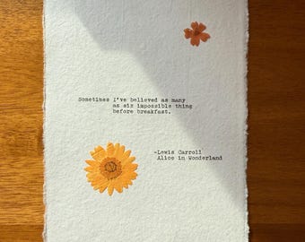 May include: A white paper print featuring a quote from Lewis Carroll's "Alice in Wonderland" and two orange pressed flowers. The quote reads, "Sometimes I've believed as many as six impossible things before breakfast."