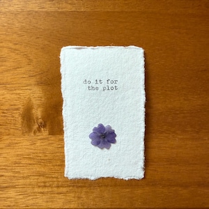 May include: A white handmade paper card with the text "do it for the plot" printed in black. A small purple flower is affixed to the card. The card rests on a wooden surface.