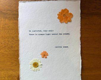 May include: A handmade paper art piece with pressed flowers and text. The white paper features an orange flower at the top, two smaller orange flowers, and a white daisy. The text reads, "Be comforted, dear soul! There is always light behind the clouds." -Little Women.