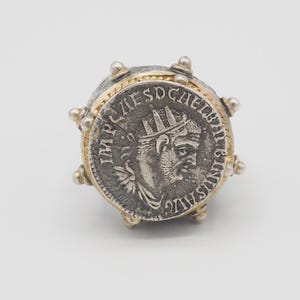 May include: A silver and gold ring featuring a coin with a profile of a man wearing a crown. The coin is surrounded by a gold band and silver accents. The ring is set against a white background.