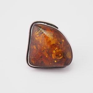 May include: A silver ring with a large, irregularly shaped amber stone. The amber is a warm, translucent brown with visible inclusions, set in a polished silver band. The ring has a modern, geometric design.