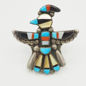 May include: A silver-toned bird-shaped brooch with inlaid turquoise, black, white, and orange stones. The bird has outstretched wings and a detailed head, showcasing a Native American design. The brooch is set against a white background.