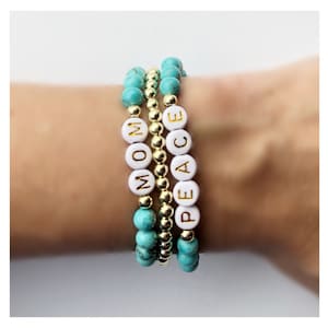 Op de afbeelding: Drie gestapelde armbanden met turquoise en gouden kralen. De bovenste armband heeft witte kralen met het woord "PEACE" gespeld in gouden letters. De middelste armband heeft gouden kralen. De onderste armband heeft witte kralen met het woord "MOM" gespeld in gouden letters.