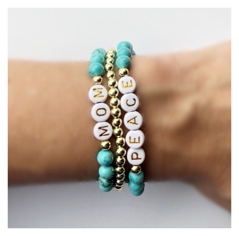 Op de afbeelding: Drie gestapelde armbanden met turquoise en gouden kralen. De bovenste armband heeft witte kralen met het woord "PEACE" gespeld in gouden letters. De middelste armband heeft gouden kralen. De onderste armband heeft witte kralen met het woord "MOM" gespeld in gouden letters.