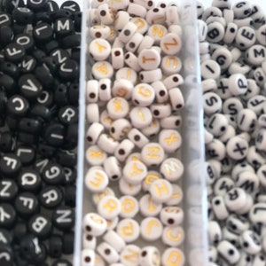 Op de afbeelding: Drie bakjes met letter kralen. Het eerste bakje heeft zwarte kralen met witte letters. Het tweede bakje heeft witte kralen met gouden letters. Het derde bakje heeft witte kralen met zwarte letters.