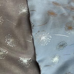 May include: Two pieces of fabric with a dandelion print. One piece is a light blue color and the other is a light brown color. Both fabrics have a silver metallic dandelion print.