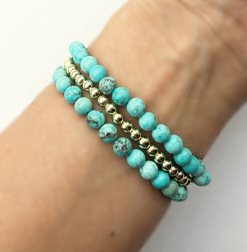 Op de afbeelding: Drie turquoise kralen armbanden met gouden kralen ertussen. De armbanden zijn gestapeld op de pols van een persoon.