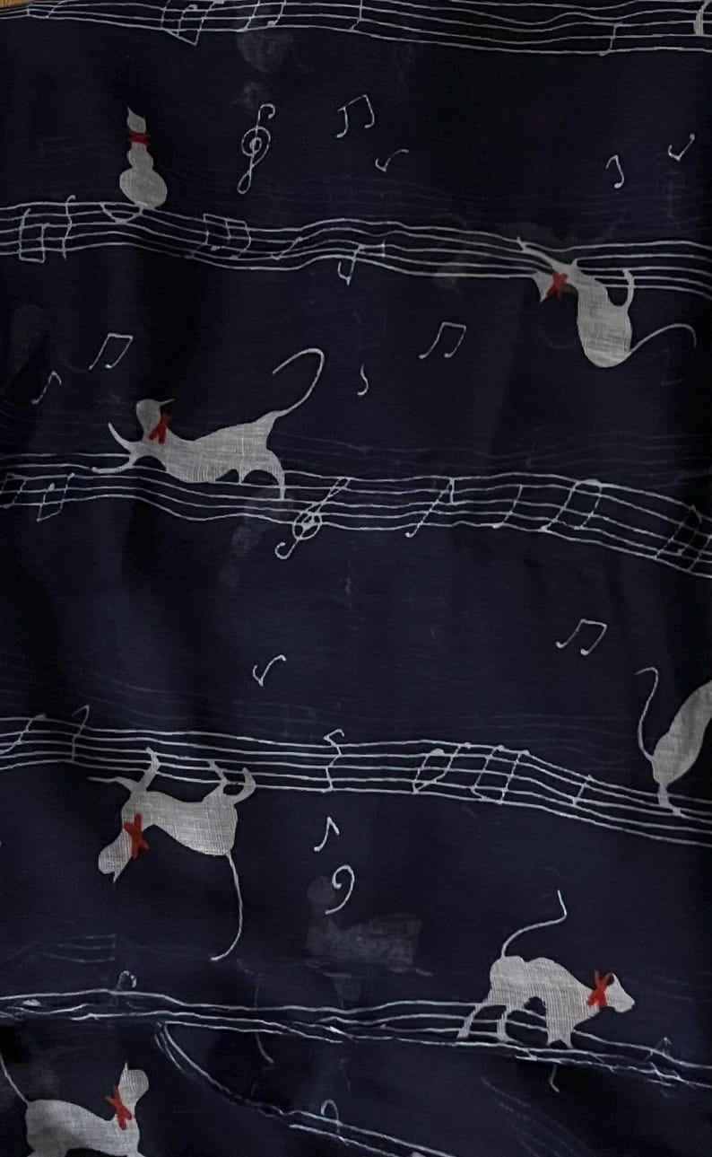 May include: A navy blue scarf with a white musical note pattern and white silhouettes of cats wearing red bows. The scarf is made of a sheer fabric.