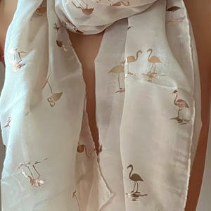 May include: A white sheer scarf with a gold flamingo print. The scarf is folded and draped around a person's neck.