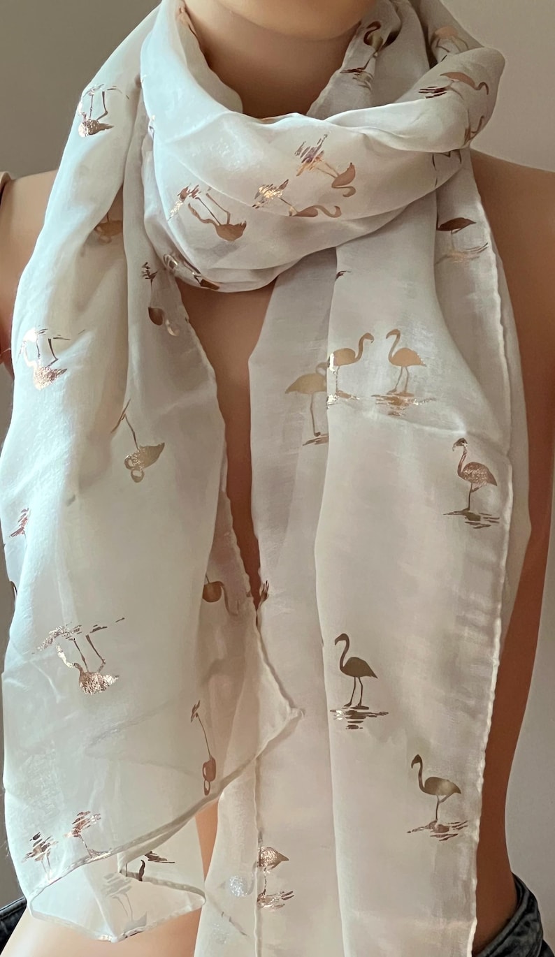 May include: A white sheer scarf with a gold flamingo print. The scarf is folded and draped around a person's neck.