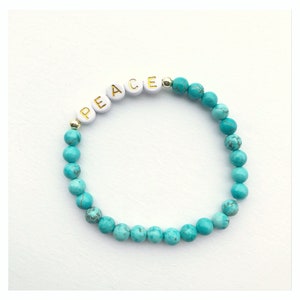 Op de afbeelding: Een turquoise kralen armband met witte letterkralen die het woord "VREDE" spellen.