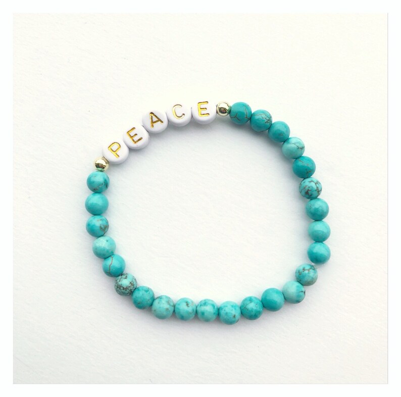 Op de afbeelding: Een turquoise kralen armband met witte letterkralen die het woord "VREDE" spellen.
