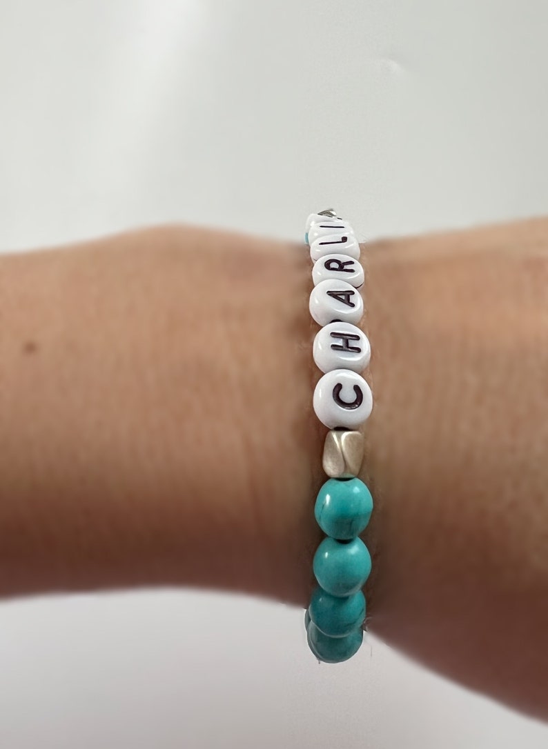 Op de afbeelding: Een turquoise en witte kralen armband met de naam "CHARLIE" gespeld in witte kralen.