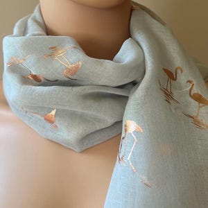 May include: A light gray scarf with a gold flamingo print. The scarf is made of a soft, lightweight fabric and is perfect for adding a touch of style to any outfit.