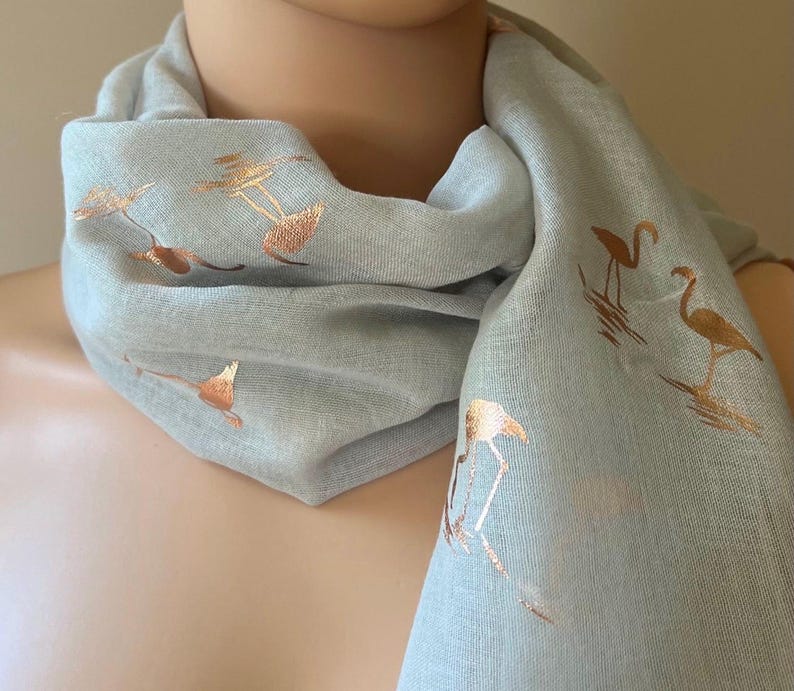 May include: A light gray scarf with a gold flamingo print. The scarf is made of a soft, lightweight fabric and is perfect for adding a touch of style to any outfit.