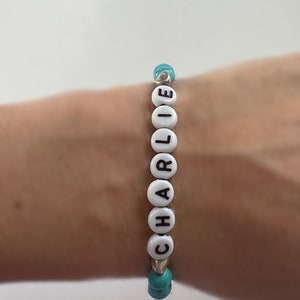 Op de afbeelding: Een witte kralen armband met de naam "CHARLIE" gespeld in zwarte letters. De kralen worden gescheiden door turquoise kralen.